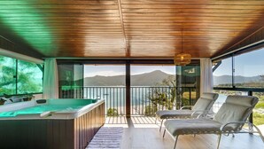 Espaço moderno em ambientes internos com hidromassagem, poltronas de descanso e vista para uma montanha e praia através de grandes portas de vidro.
