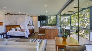 Quarto moderno com uma cama grande, uma escrivaninha de madeira e vista para árvores através de janelas amplas.