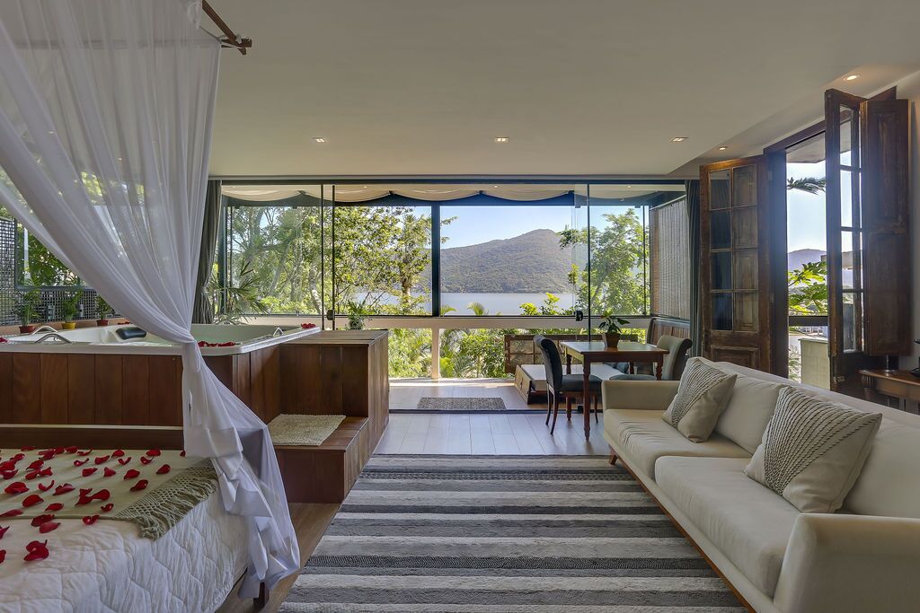 A bedroom with a large window, a bed with rose petals, a sitting area with a sofa and chairs, and a view of a lake and mountains.