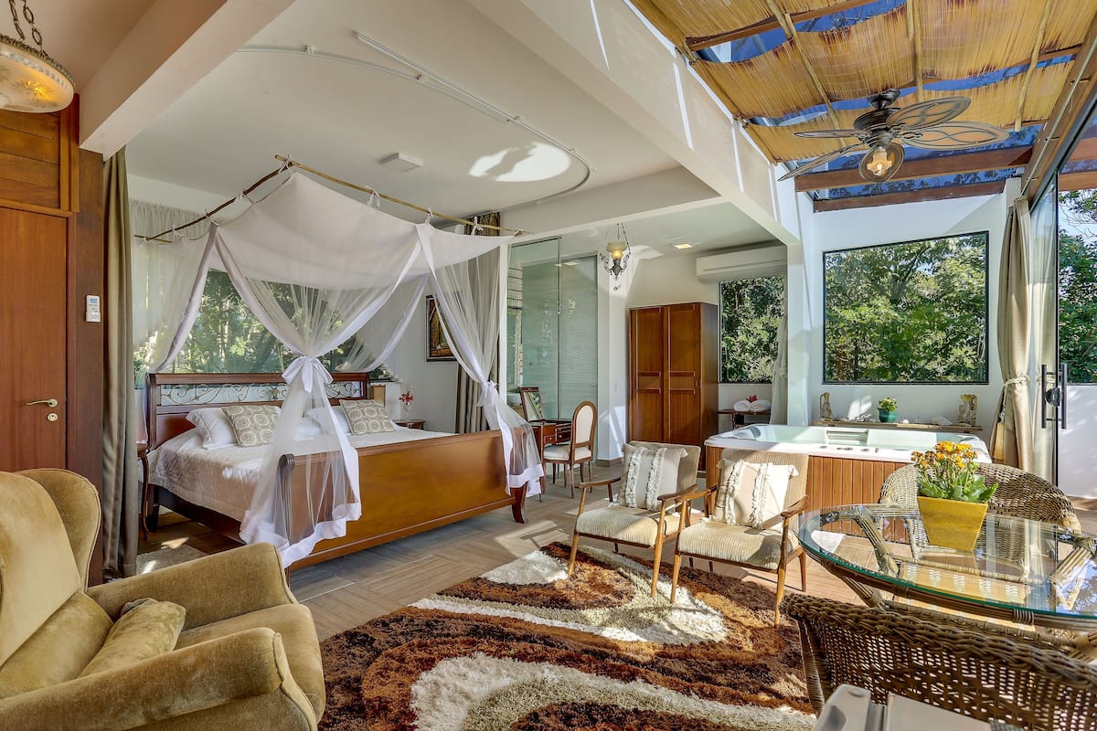 Un dormitorio con cama con dosel, una zona de estar con sillas, una mesa de vidrio y vistas al exterior.
