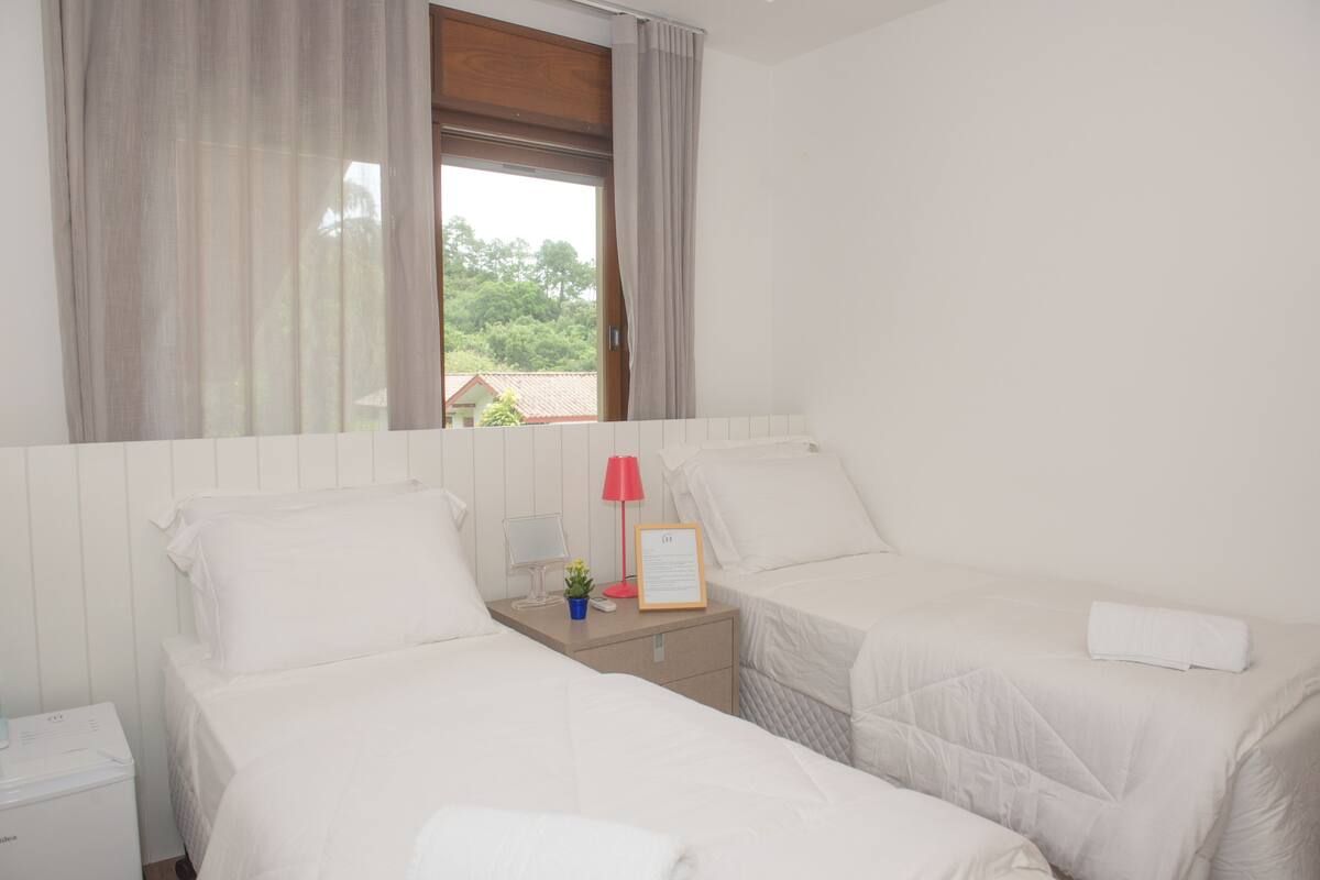 A hotel room with two beds, a nightstand with a lamp, a small plant, and a window with curtains.