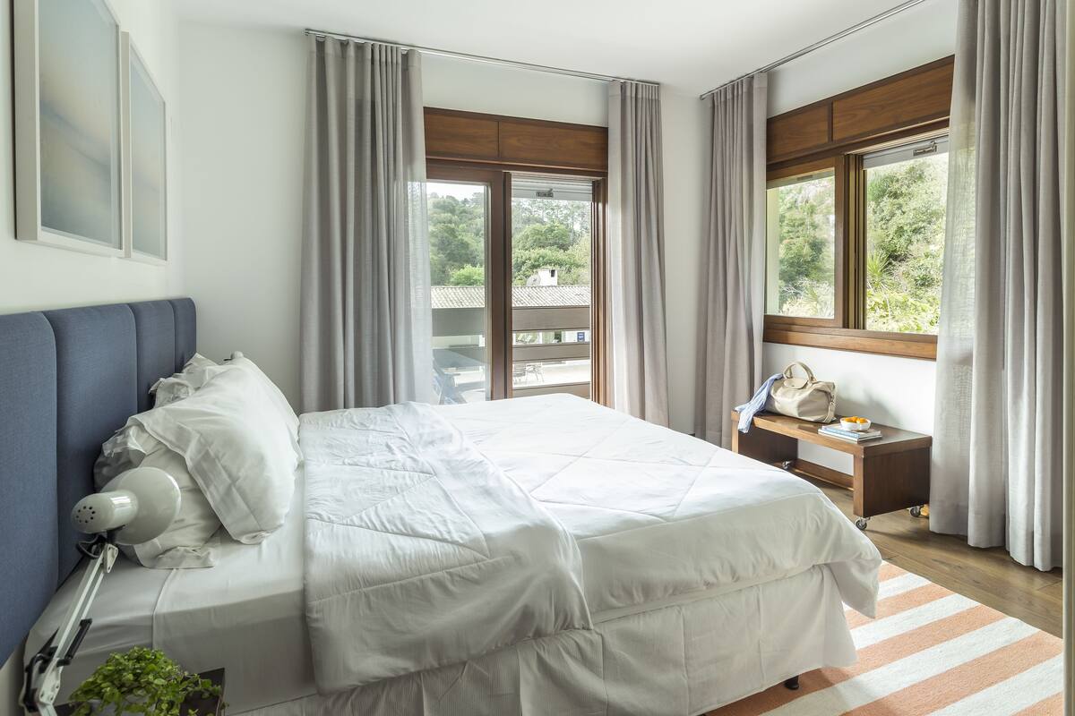 A neatly made bed with white linens, a bedside table with a lamp, and a view of greenery outside.