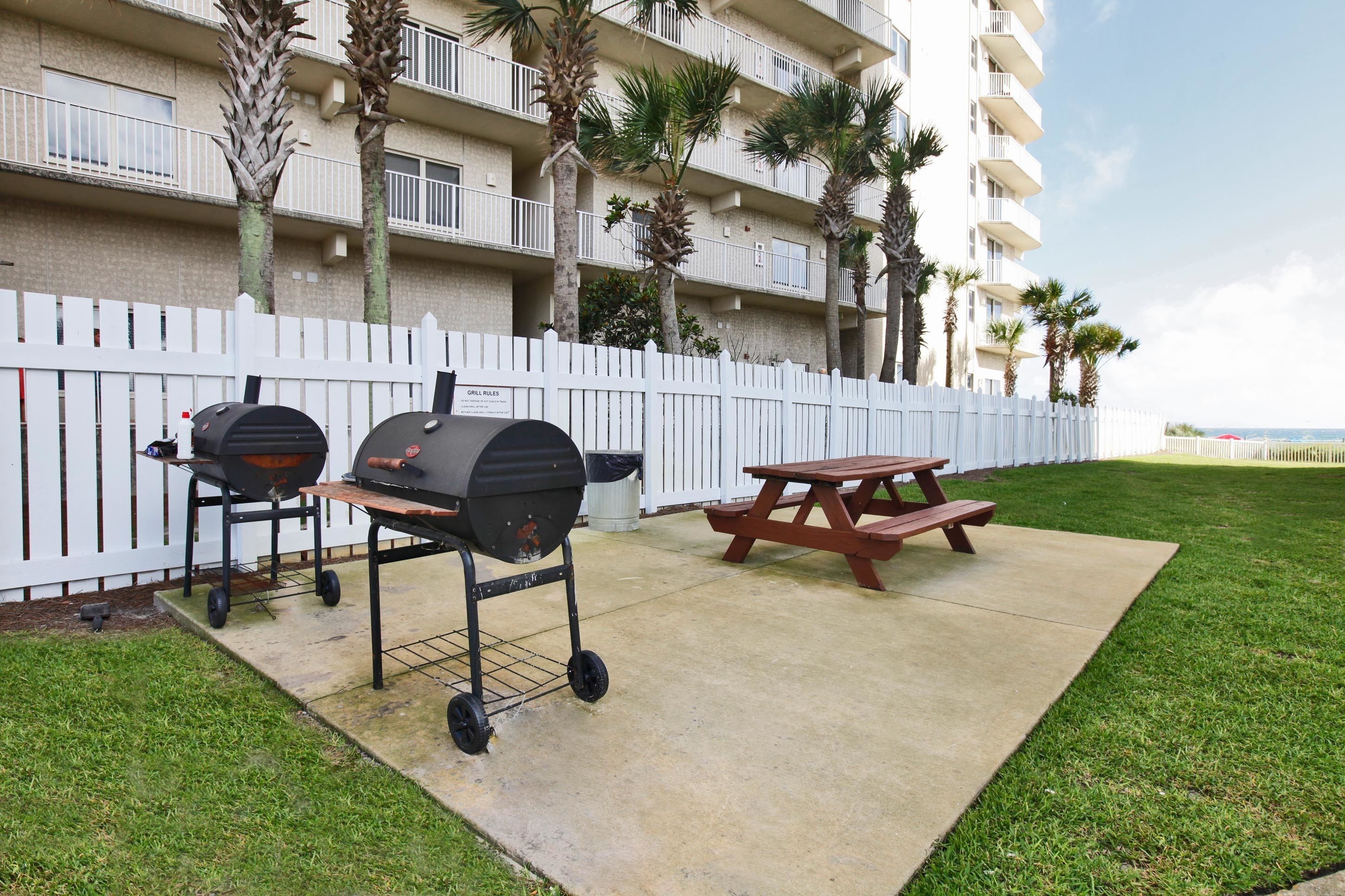 An outdoor patio area with a grill and picnic table, surrounded by a white fence and palm trees.