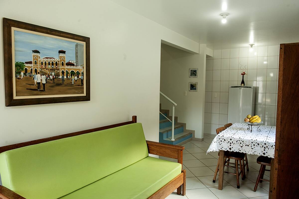 A room with a green sofa, a dining table with a lace tablecloth, a refrigerator, and a staircase.