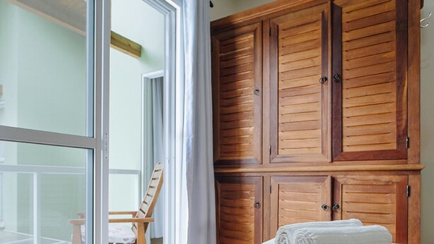 Habitación de hotel con cama, armario de madera y una silla junto a un ventanal corredero.