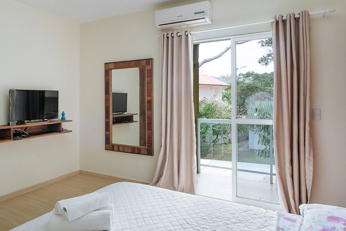 A bedroom with a bed, a TV on a shelf, an air conditioner, and a balcony with curtains.