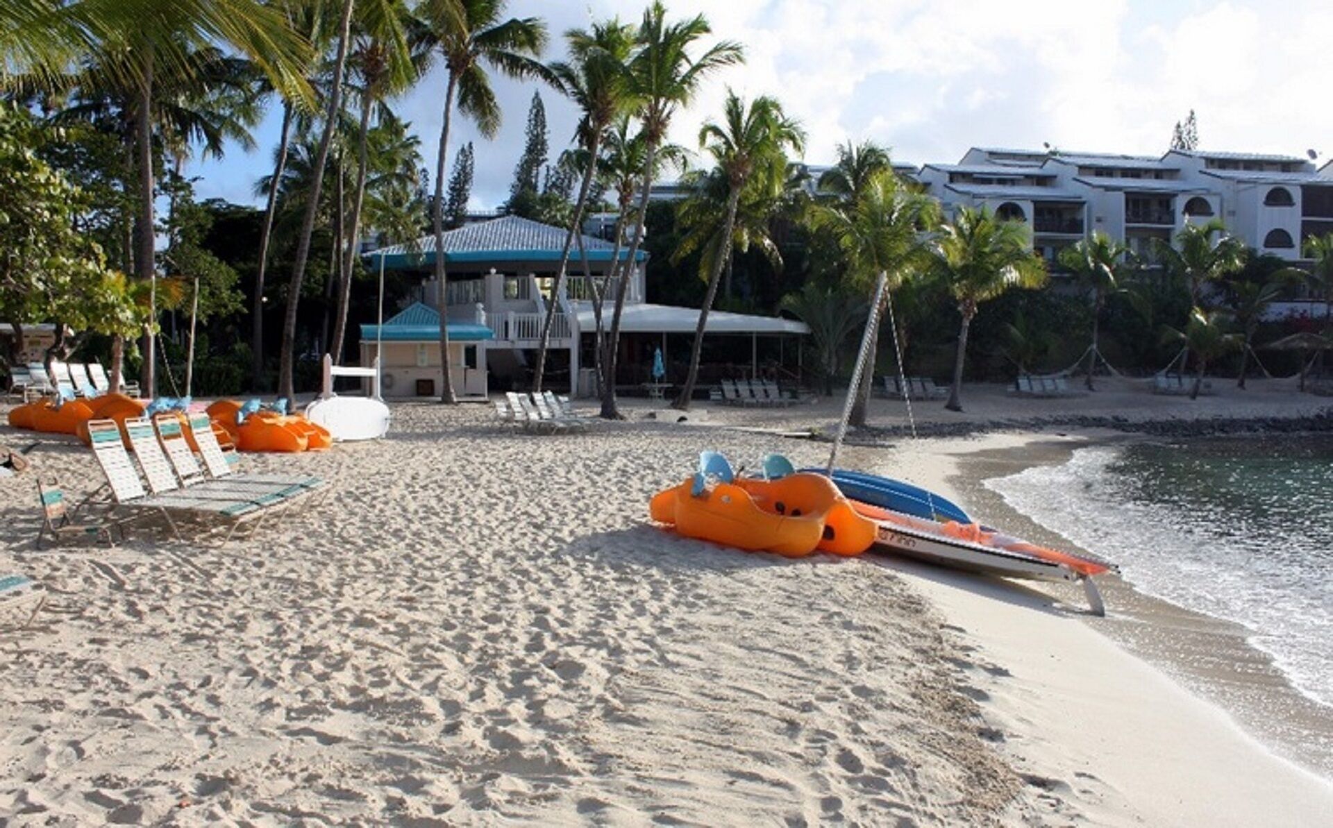 A beach with lounge chairs, a pool, and a building with a blue roof.