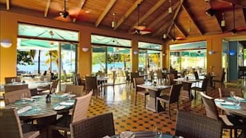 Un comedor amplio con techos altos y ventanas grandes, que presenta un suelo de cuadros y mobiliario de mimbre.
