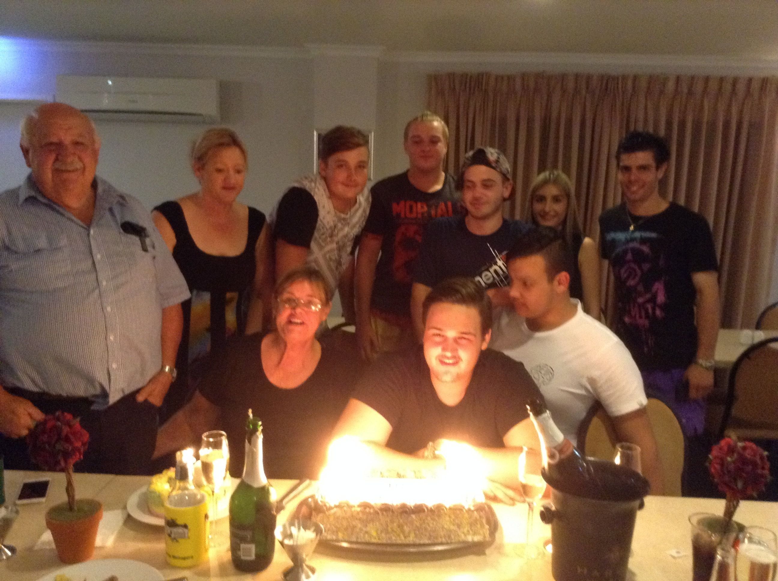 A group of people gathered around a table with a lit birthday cake, champagne bottles, and a potted plant.