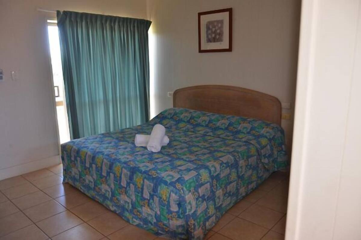 A single bed with a patterned bedspread and rolled towels, a wooden headboard, a framed picture on the wall, and a window with curtains.