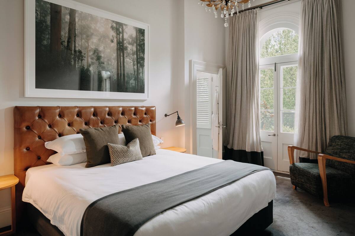 A bedroom with a large bed, a framed forest painting, a chandelier, and a view of the outdoors through a window with curtains.