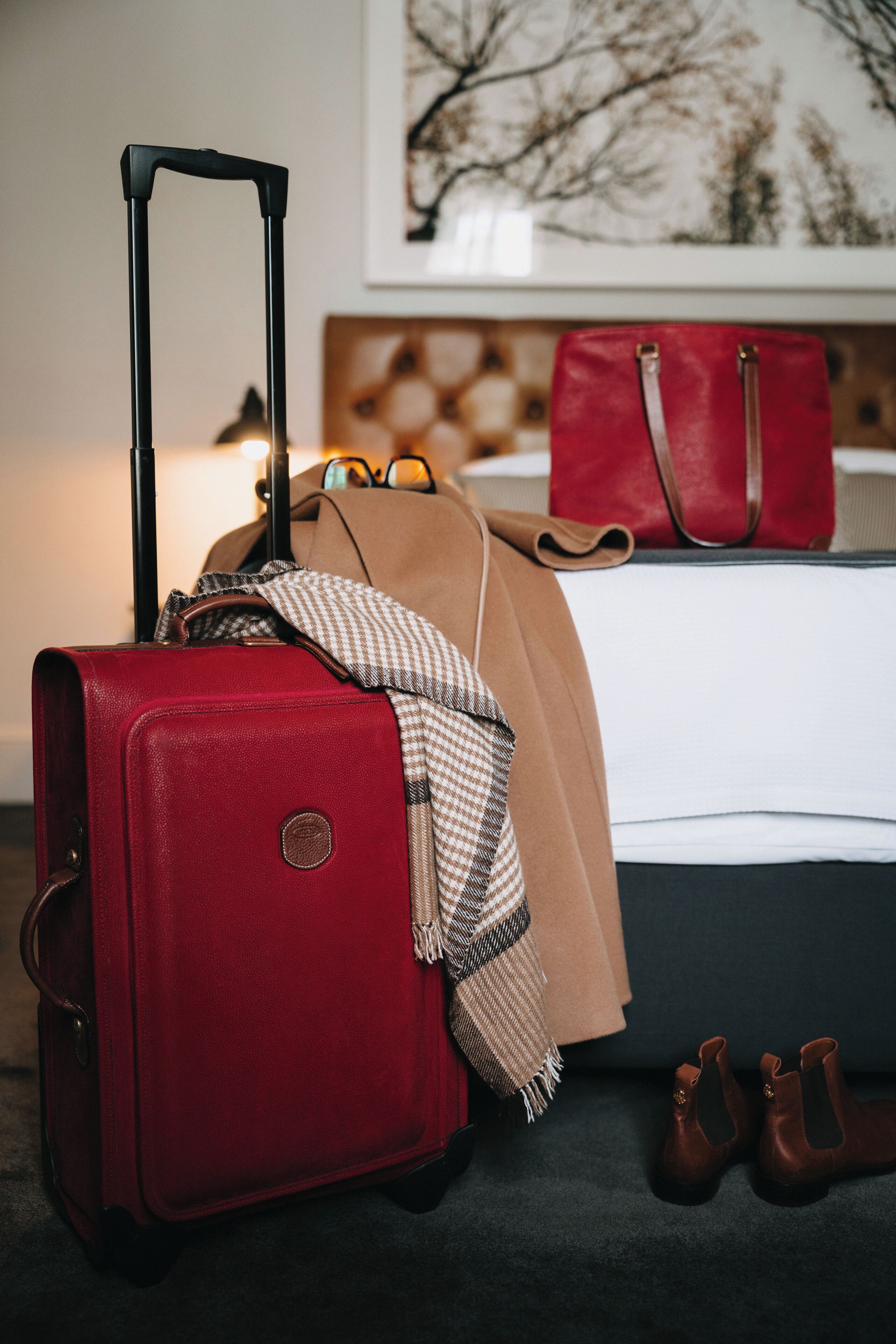 A room with a bed, a red suitcase, a brown coat, and a pair of brown boots.