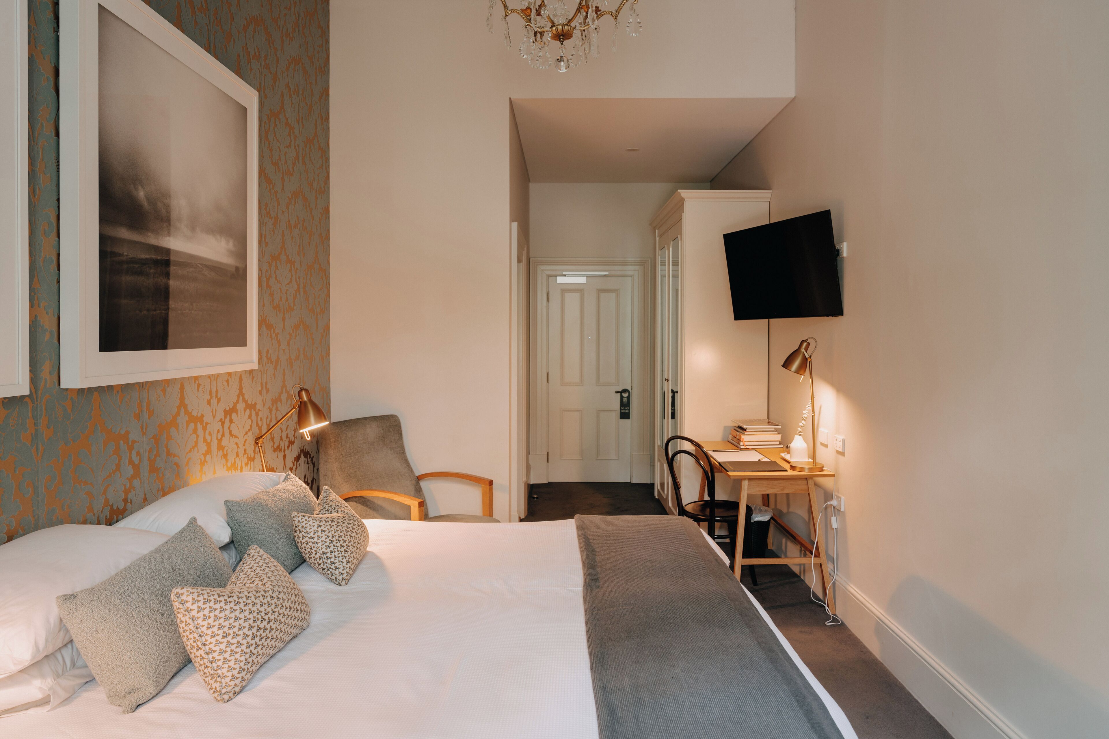 A hotel room with a bed, pillows, a desk with a chair, a television, and a chandelier.