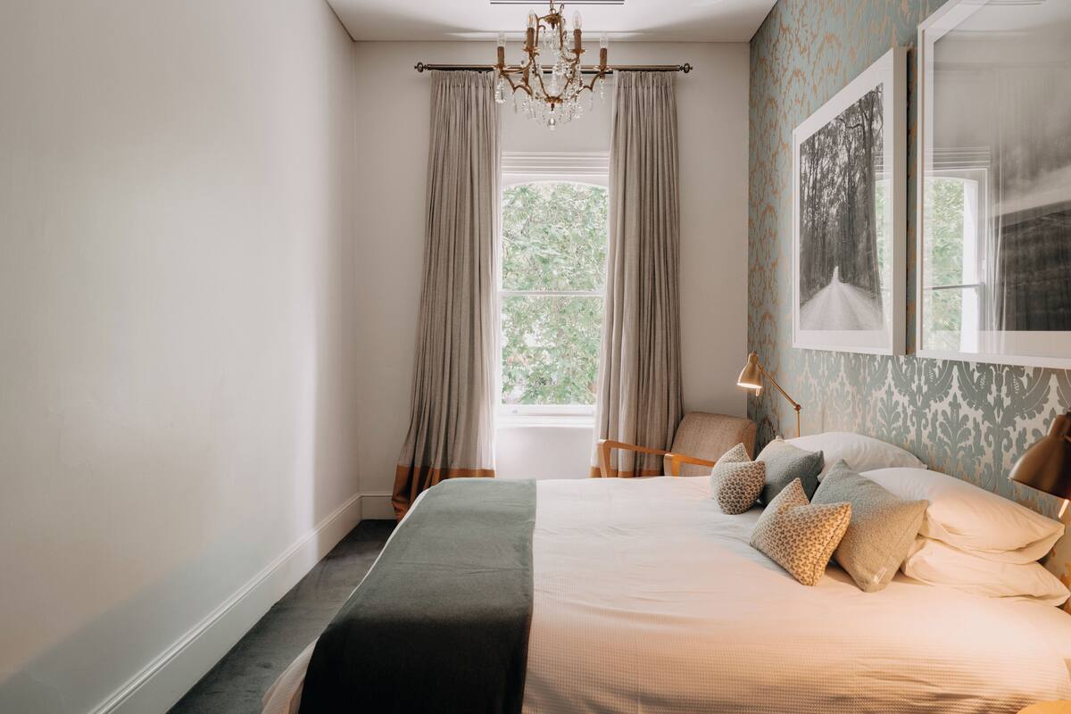 A bedroom with a large bed, a window with curtains, a chandelier, and a framed picture on the wall.