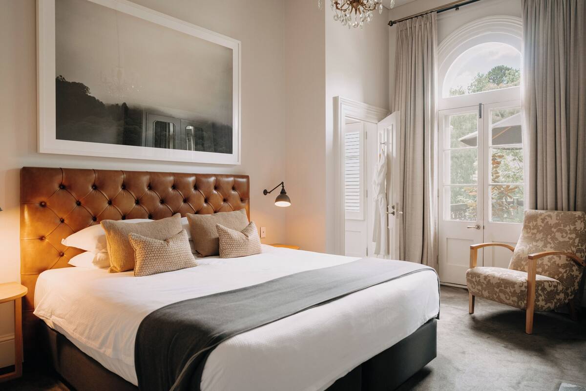 A bedroom with a large bed, a headboard, a framed picture on the wall, a chair, and a window with curtains.