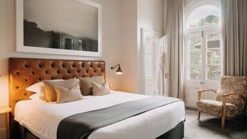 Un dormitorio con una cama grande, cabecera, un cuadro enmarcado en la pared, una silla y una ventana con cortinas.