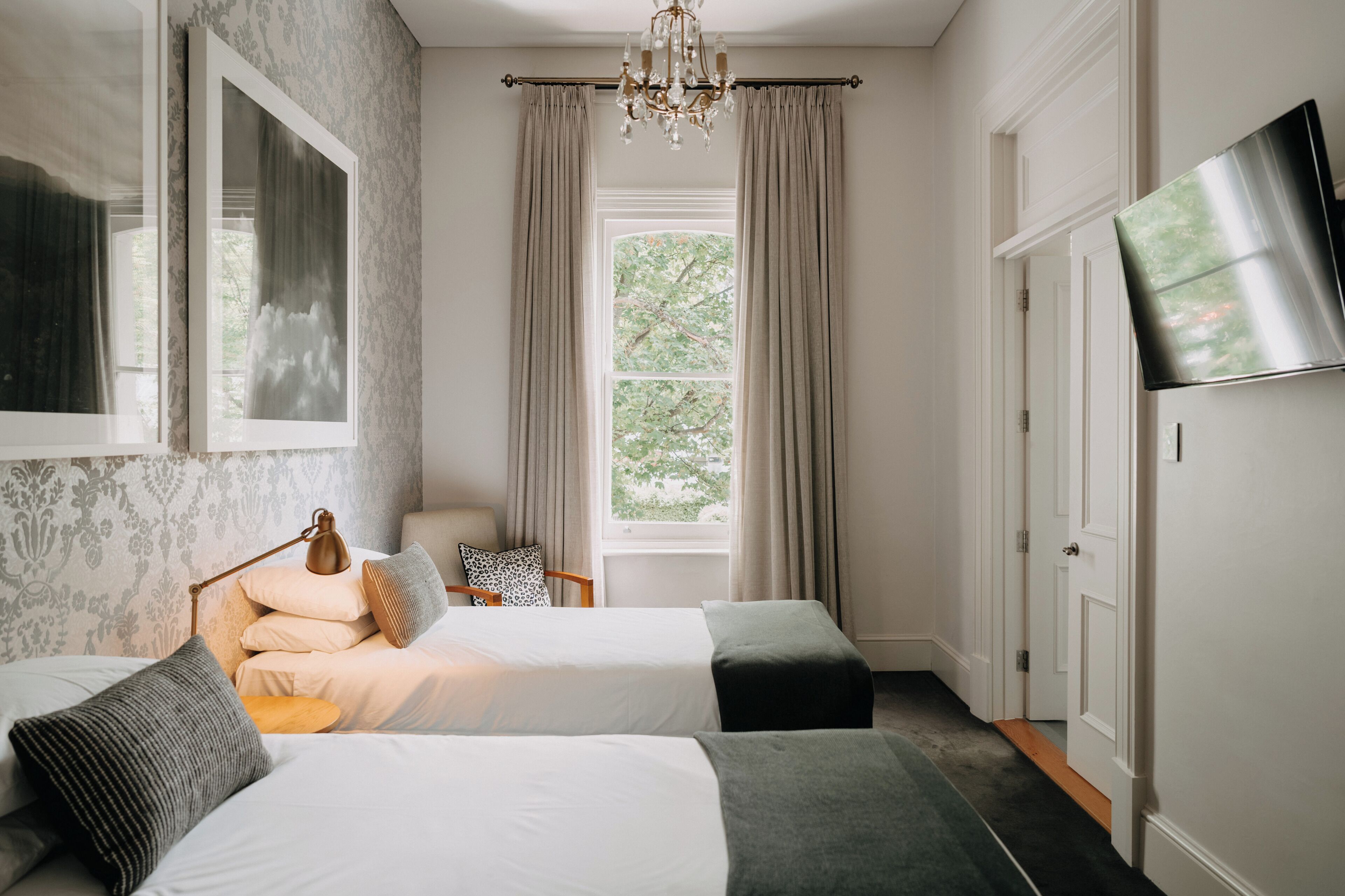 A bedroom with two beds, a chandelier, a TV, and a window with curtains.