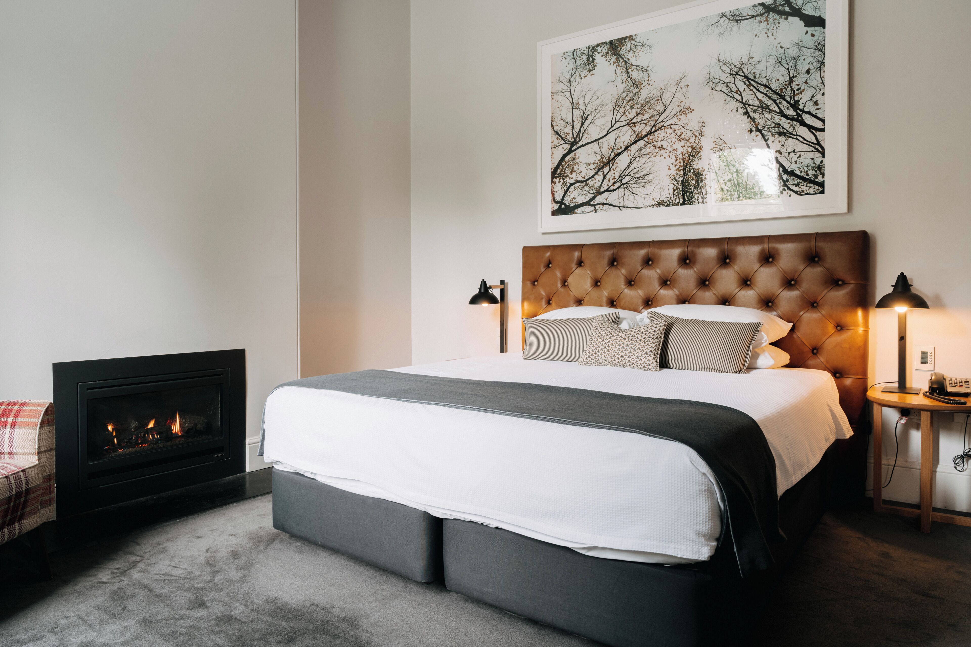 A bedroom with a fireplace, a large bed with a tufted headboard, bedside lamps, and a framed picture on the wall.