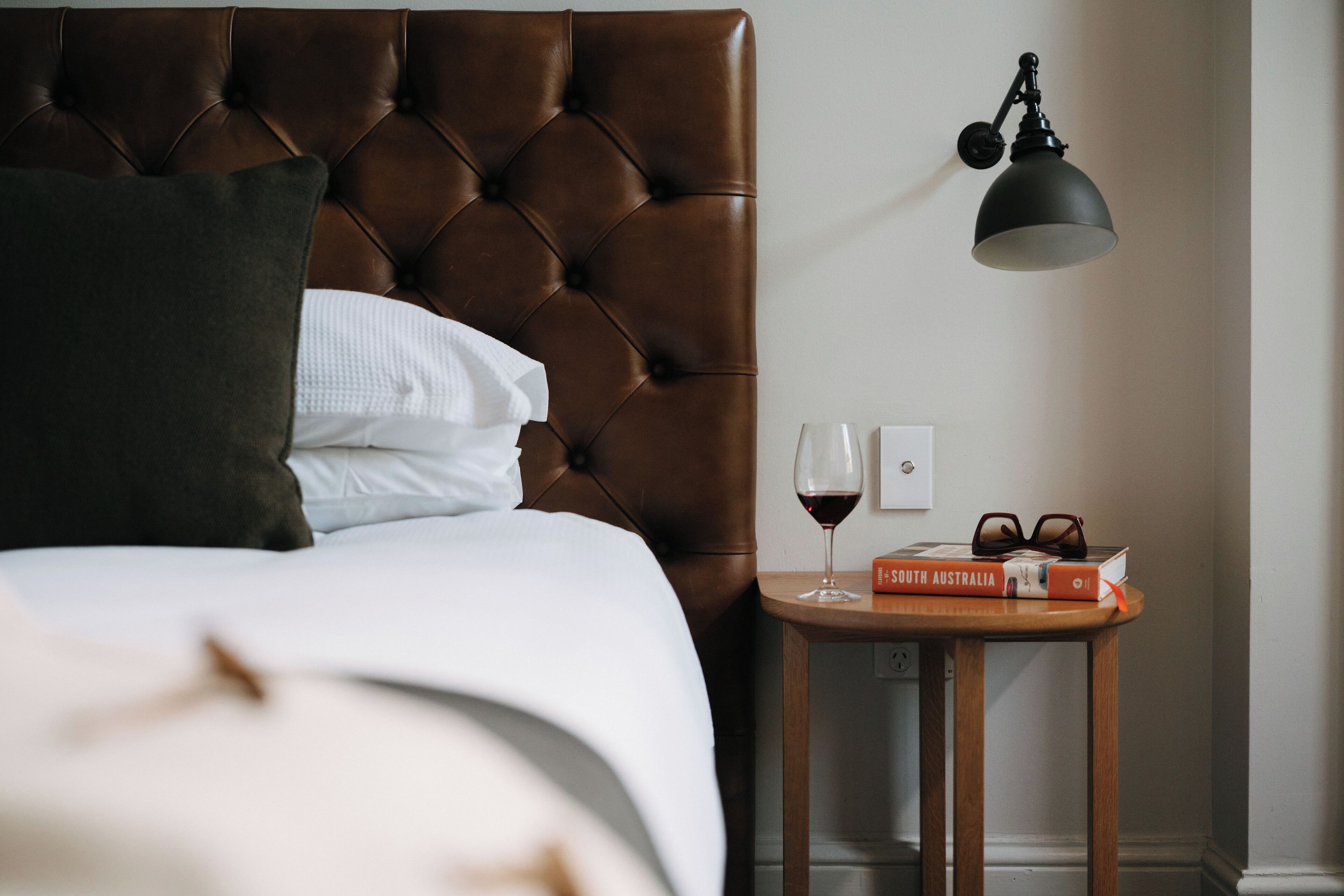 A neatly made bed with a brown headboard, a bedside table with a book and a wine glass, and a wall-mounted lamp.