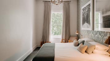 A bedroom with a large bed, a chandelier, a window with curtains, and a wall with a framed picture.