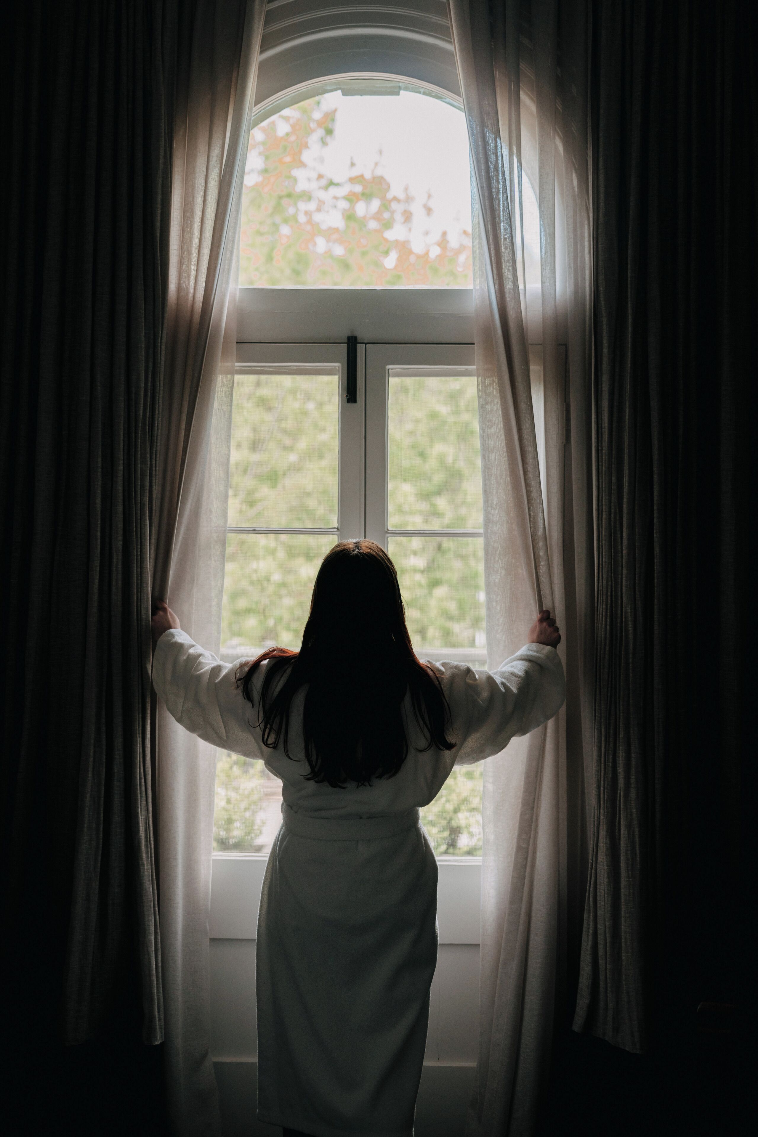 A person standing in front of a window with curtains, looking outside.