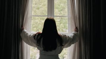 A person standing in front of a window with curtains, looking outside.