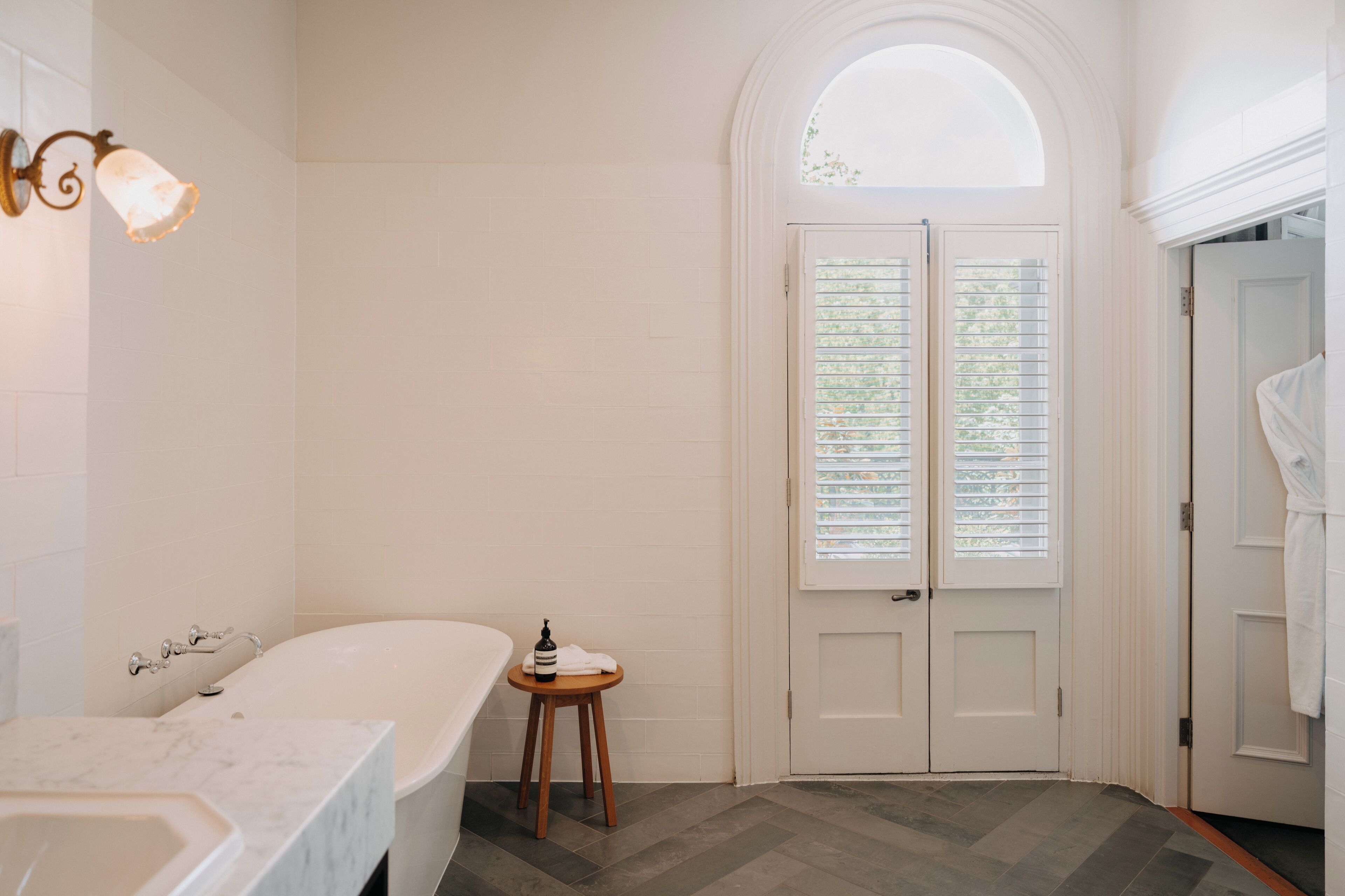 Une salle de bain avec une baignoire, un lavabo et un meuble-lavabo. On y trouve un tabouret en bois sur lequel sont posés une bouteille et une serviette. Le sol est recouvert de parquet disposé en point de Hongrie, et la porte est blanche, avec une fenêtre au-dessus.