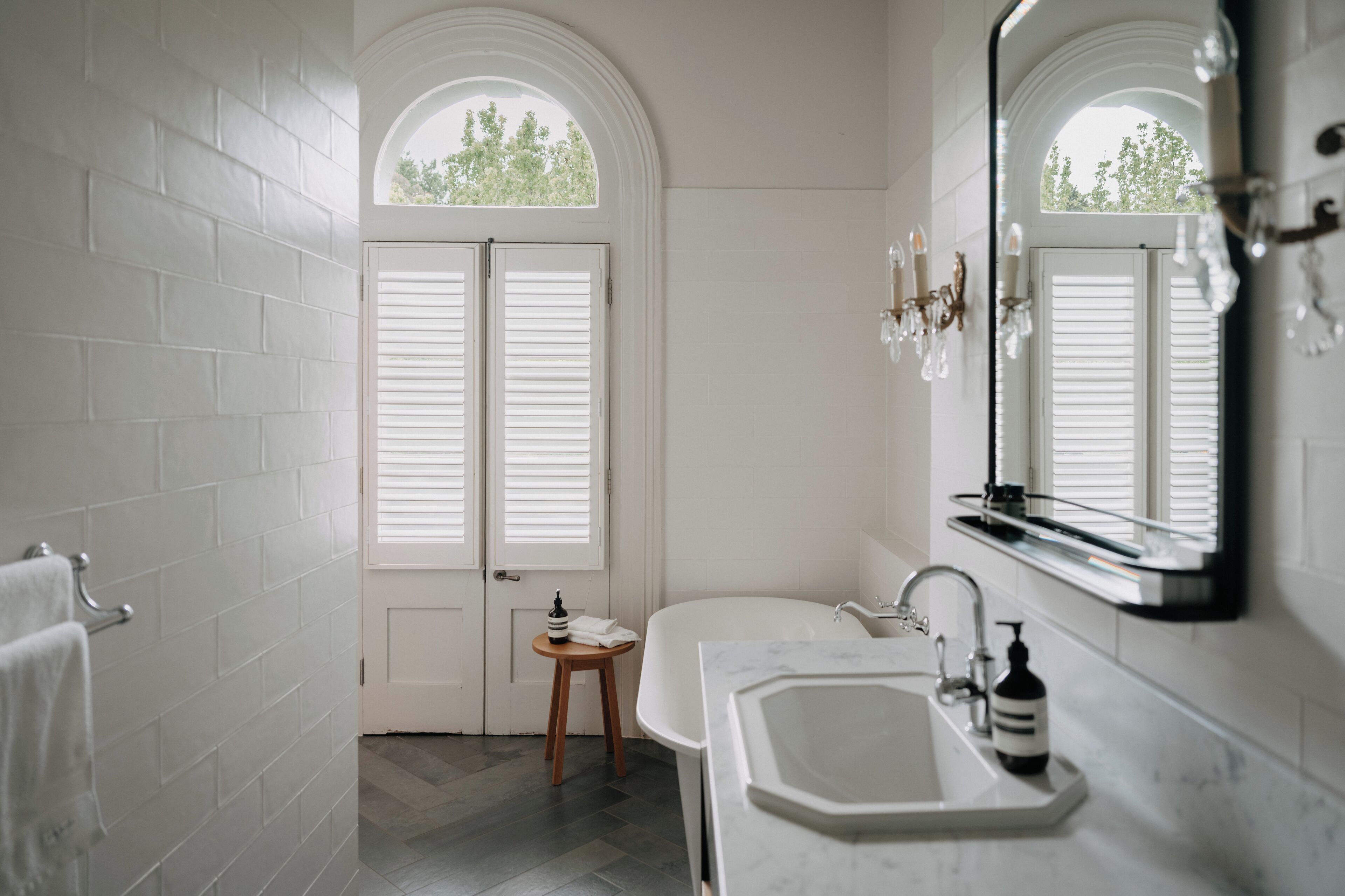 A bathroom with white brick walls, a white bathtub, a sink with a mirror, and a small wooden stool.