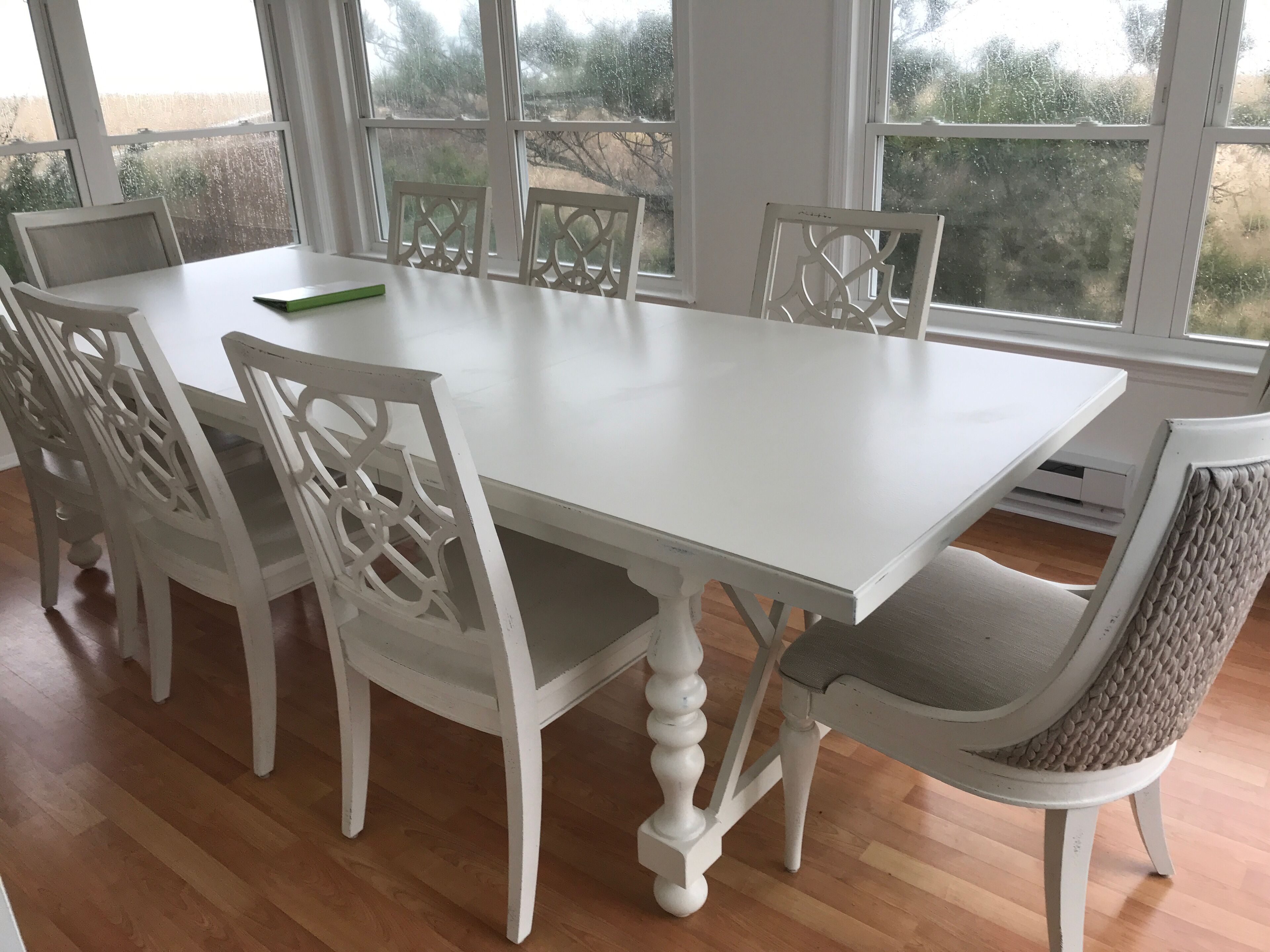 A white dining table with six chairs, large windows with a view of trees and a stone wall, and a wooden floor.
