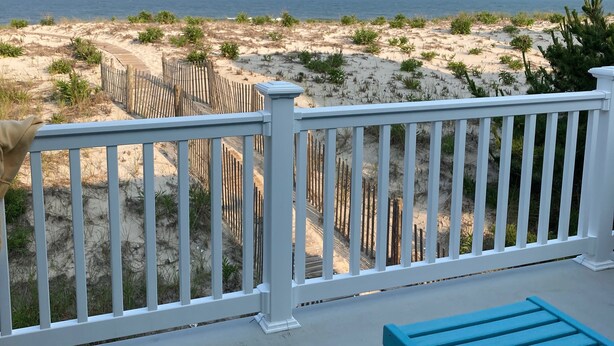 A beach view from a balcony with a wooden fence, sandy beach, and two blue lounge chairs.