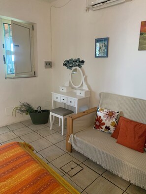 Ein Zimmer mit einem Bett, einer weißen Kommode mit Spiegel, einem Sofa mit einem Blumenkissen, einer Topfpflanze und einem Fenster.