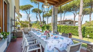 Un'area pranzo all'aperto con un tavolo apparecchiato per un pasto, circondato da sedie e con vista su un giardino con alberi e un edificio sullo sfondo.
