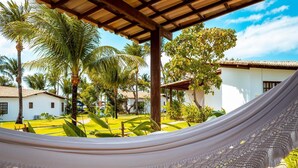 A hammock in a lush garden with palm trees and a white building in the background. - Acquapipa Resort (Tibau do Sul)