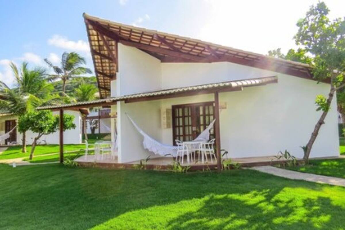 A single-story building with a white exterior and a sloped roof. There is a covered patio area with a hammock and a table.