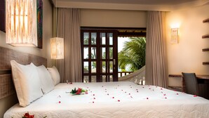 A hotel room with a large bed, a wooden headboard, a nightstand with a vase of flowers, a desk with a chair, and a view of palm trees through a window. - Acquapipa Resort (Tibau do Sul)