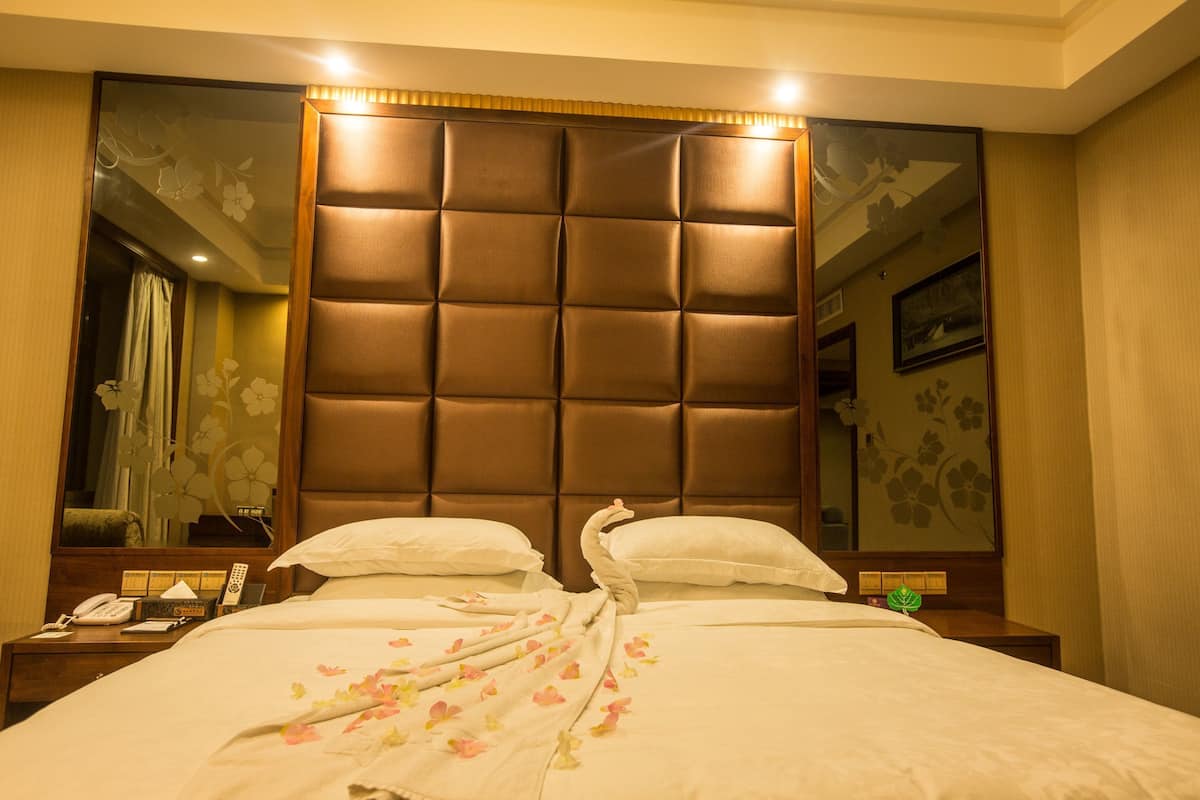 Une chambre d’hôtel avec un grand lit, une tête de lit en bois, une table de chevet avec un téléphone et une lampe, et un miroir au mur.
