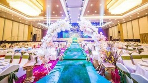Un salón de banquetes con filas de sillas blancas, un pasillo central decorado con flores y un escenario al fondo.