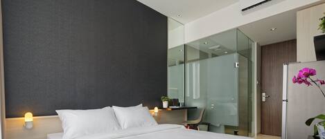 A modern hotel room with a large bed, bedside lamps, a desk with a chair, a glass partition, and a small plant.