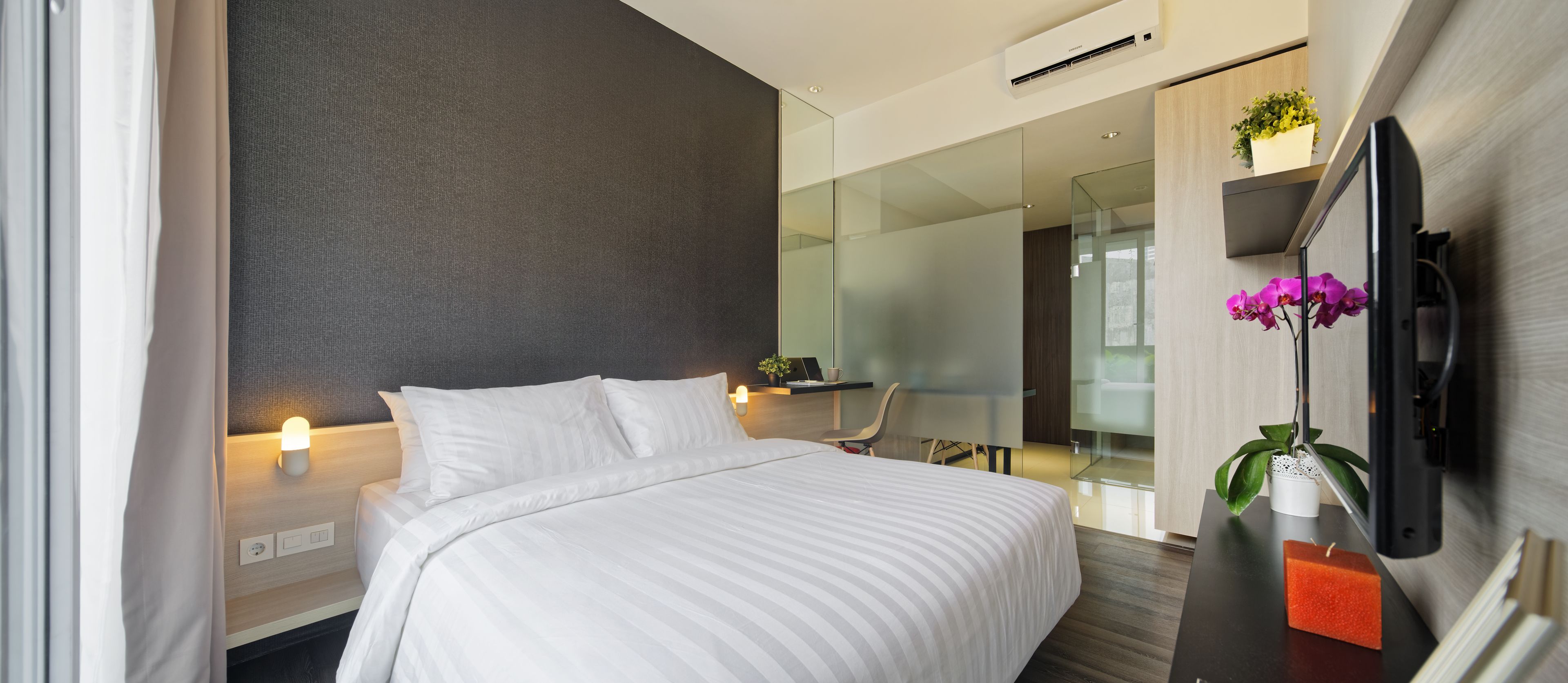 A modern hotel room with a bed, bedside table, TV, and a glass partition separating the room from a dining area.