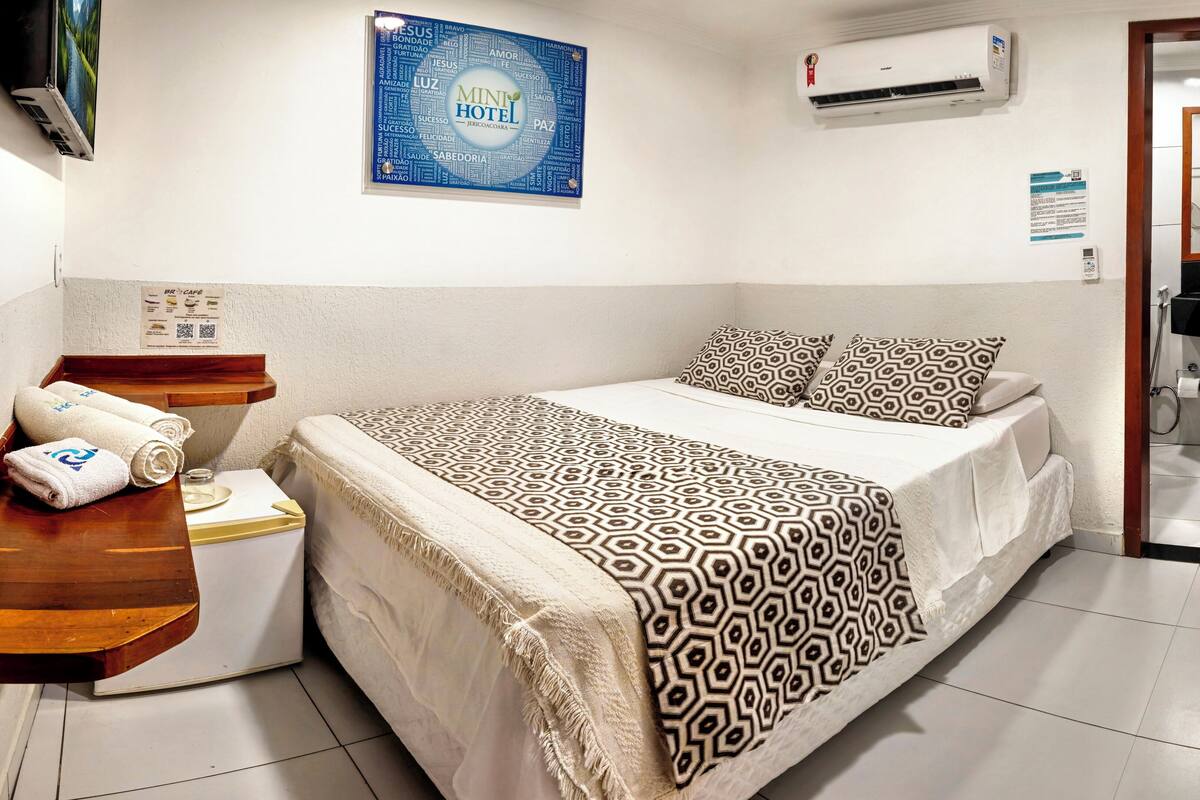 Une chambre d’hôtel avec un lit, une table de chevet avec des serviettes, un climatiseur fixé au mur et une porte de salle de bain visible.