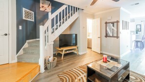 A modern living room with a staircase, a flat-screen TV, a ceiling fan, and a glass coffee table.