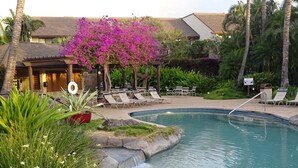 Piscina com espreguiçadeiras e área de estar coberta, cercada por plantas tropicais e um edifício ao fundo.