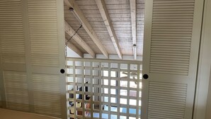 Quarto com teto de madeira, persianas brancas e vista através de uma janela com treliça.