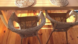 Amplia habitación con techo alto, piso de madera y una gran mesa con individuales tejidos. Alrededor de la mesa hay sillas de mimbre, y se ve el océano a través de amplias ventanas.