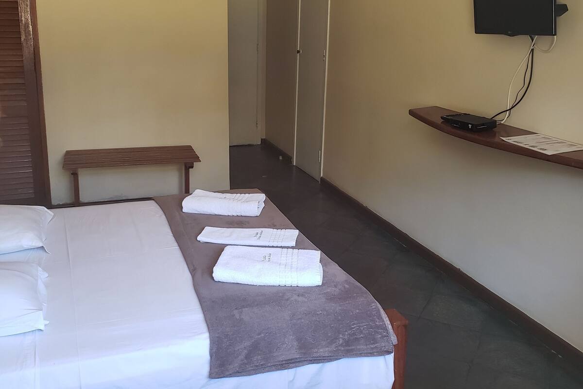 A hotel room with a bed, a bench, a TV mounted on the wall, and a small table with a power outlet.