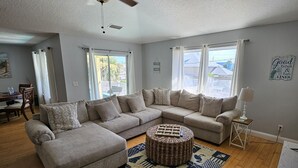 A spacious living room with a large L-shaped sectional sofa, a round ottoman, and a ceiling fan. - 1ST STREET II BEACH HOUSE, UPDATED DOG FRIENDLY  FENCED IN YARD 1 BLOCK TO BEACH (St. Augustine)