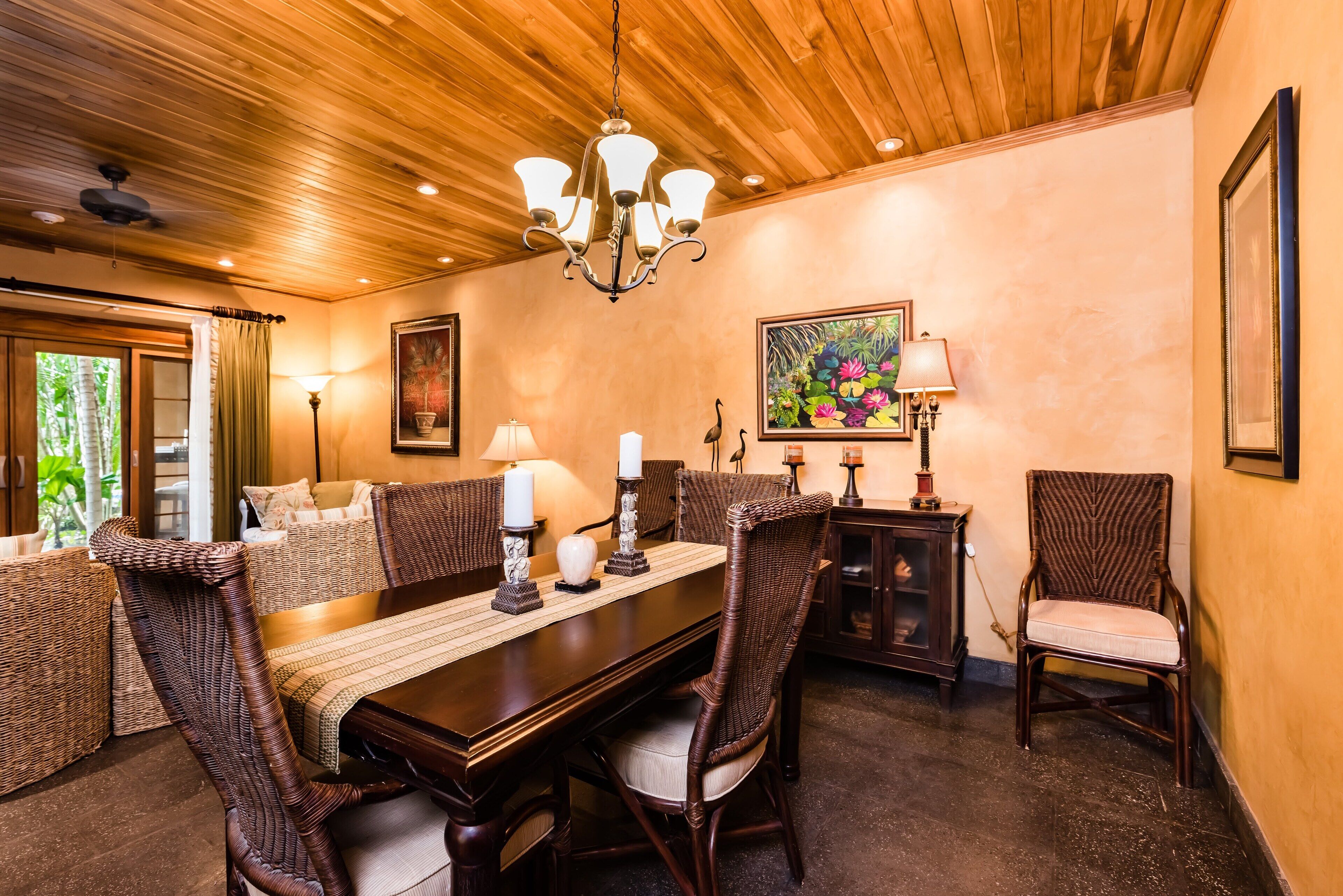 A dining area with a wooden ceiling, a chandelier, wicker furniture, and framed artwork on the walls.