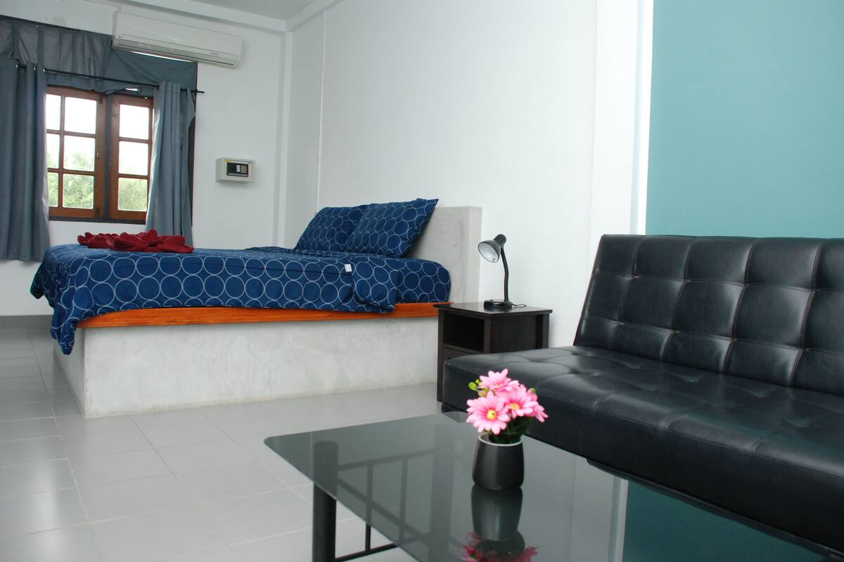 A bedroom with a bed, a black leather sofa, a small table with a vase of flowers, a nightstand, and a window with curtains.