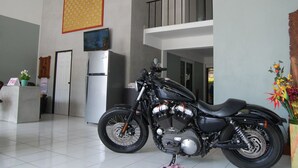 A motorcycle in a spacious, modern living area with a balcony, a refrigerator, and a decorative plant. - Sea & Sand Guesthouse and Restaurant (Cha-am)
