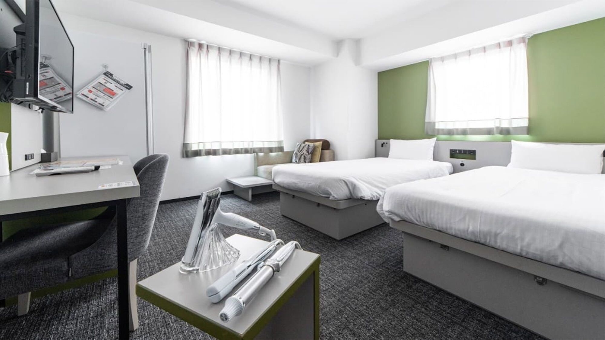 Hotellihuone, jossa on työpöytä, kaksi sänkyä, tuoli, pieni pöytä, jossa on puhelin ja lasi, sekä ikkuna, jossa on verhot.